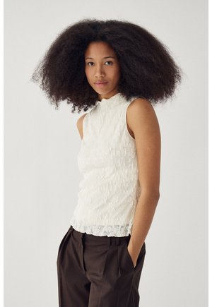 Giovane donna con capelli ricci e voluminosi, che indossa un top bianco senza maniche e pantaloni marroni scuri, in piedi con una mano nella tasca.