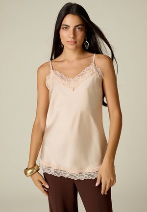 Donna con lunghi capelli scuri indossa una canottiera beige con rifiniture in pizzo, pantaloni scuri, orecchini geometrici e un grande braccialetto d'oro al polso sinistro.