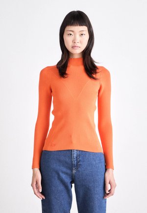 Pull en rib à col montant orange, avec des manches longues ajustées et un motif en V sur le devant. Associé à un jean en denim bleu.