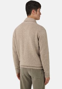 Man som står med ryggen mot kameran, iklädd en beigemelerad stickad tröja med ribbade muddar och olivgröna kordbyxor mot en vit bakgrund.