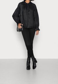 Svart långärmad blus med rund krage, texturerad tyg och spetskant. I kombination med svarta leggings och ankelstövlar, bärande på en liten väska.