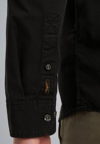 Schwarzes Langarmshirt mit Knopfmanschette, strukturiertem Stoff und einem orangefarbenen Logopatch in der Nähe der Naht. Stilvolles, figurbetontes Design.