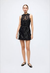 Black sleeveless dress with a high neck, featuring a sheer overlay and decorative patterns, cinched waist, and flared skirt.