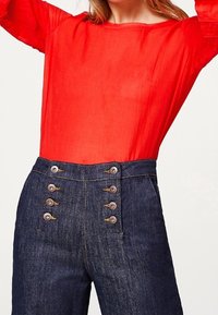 Rød, let langærmet top parret med høj talje mørkeblå denim bukser med seks knapdetaljer foran.