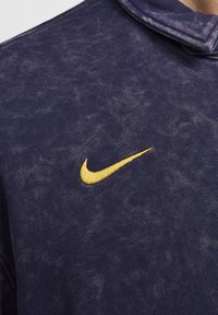 Navy sininen huppari, jossa on teksturoitu pinta ja keltainen brodeerattu Nike-logo rinnassa. Varustettu klassisella hupulla ja ribbailla hihoilla.