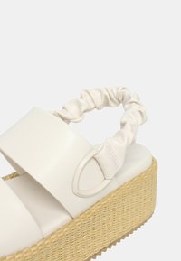 I sandali platform bianchi presentano una base in giute intrecciato e chunky, una tomaia morbida e lucida e una fascia arricciata. Accento distintivo in metallo ovale sul lato.