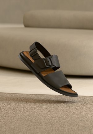 Schwarze Sandale mit zwei verstellbaren Riemen, Obermaterial aus glattem Kunstleder, gepolsterte braune Innensohle, flache Gummisohle und offenem Zehendesign.