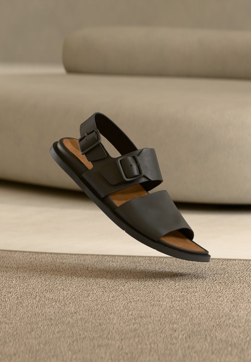 Μαύρο σανδάλι με δύο ρυθμιζόμενους ιμάντες, επάνω μέρος από λείο τεχνητό δέρμα, μαλακή σόλα σε καφέ χρώμα, επίπεδη σόλα από καουτσούκ και ανοιχτό σχέδιο στα δάχτυλα.