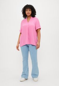 Rosa, kortermet bluse med V-hals og oppbrettede ermer, paret med lyseblå, utsvingte jeans og hvite joggesko. Glatt tekstur.