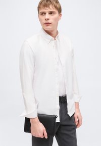 Νέος άνδρας με λευκό πουκάμισο με κουμπιά και γκρι παντελόνι, κρατώντας μαύρο πορτοφόλι με φερμουάρ, κοιτάζει προς το πλάι με φόντο απλό.