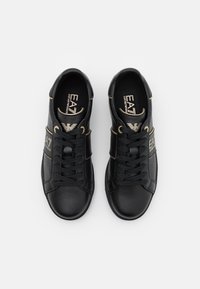 Ett par svarta EA7 Emporio Armani sneakers med snören, beige detaljer och logotyp på sidan och tungan, visade uppifrån mot en vit bakgrund.