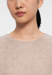 Light beige sweater with a fine knitted texture, round neckline, and ribbed trim, shown on a person, focusing on the upper chest area.