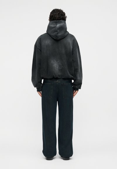 Άτομο με σγουρά μαλλιά φοράει ένα πλυμένο μαύρο φούτερ με κουκούλα και φαρδιά μαύρη παντελόνια, γυρισμένο πλάτη σε απλό φωτεινό φόντο.