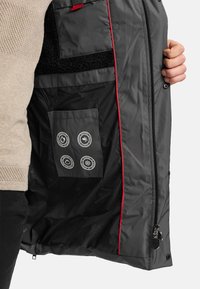 Binnenkant van een grijze jas met een zwarte fleece voering, rode accenten en een buitenzak met vier weersonderdeel-iconen. Ritssluiting zichtbaar.