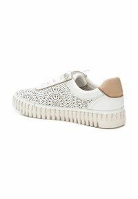 Zapatilla blanca con parte superior perforada que presenta patrones circulares, un acento en el talón de cuero marrón y una suela de goma gruesa y texturizada.