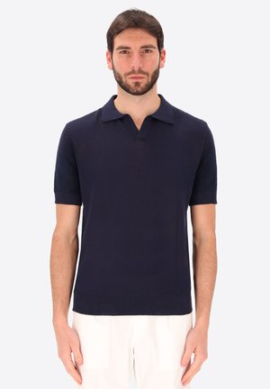 Uomo con capelli corti e scuri e barba, che indossa una polo blu navy tinta unita a maniche corte e pantaloni chiari, in piedi su uno sfondo bianco.