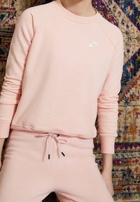 Ljusrosa sweatshirt och matchande byxor i mjukt tyg, med rund halsringning och raglanärmar. Sweatshirten har en liten vit logotyp.