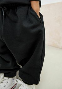 Die Hand eines Kindes steckt in der Tasche eines schwarzen Sweatpants, das Kind trägt weiße Schnürsenkel-Sneaker und steht auf einem hellen Boden vor einer beigen Wand.