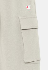 Lys grå cargo-bukser laget av teksturert stoff, med en sidelomme og en liten brodert logo i rødt og hvitt på den øvre delen av benet.