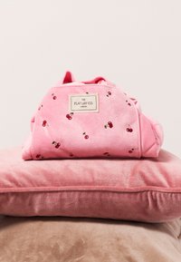 Bolso de peluche rosa con bordado de cereza, de forma rectangular y con una etiqueta de la marca. Colocado sobre suaves cojines en tonos rosa y beige.