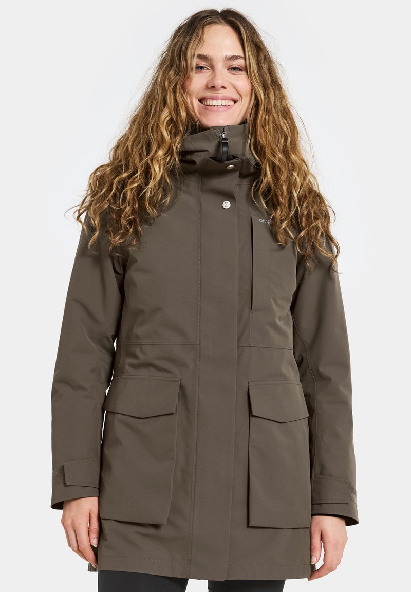Kobieta uśmiechająca się, ubrana w wodoodporną kurtkę w kolorze oliwkowym o długości do kolan, z dużymi kieszeniami na klapach i wysokim kołnierzem, stojąca na tle białym.