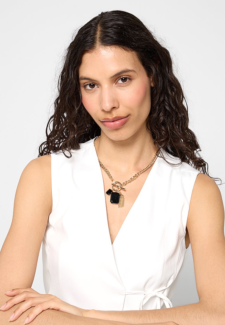 Złoty naszyjnik z łańcuszka z czarnymi geometrycznymi zawieszkami i akcentem w postaci frędzla, prezentowany na białej bezrękawnikowej bluzce.