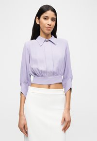 Γυναίκα που φοράει ένα κοντό λιλά μπλούζα με φαρδύ γιακά και μανίκια τριών τετάρτων, συνδυασμένη με μια λευκή φούστα ψηλής μέσης, μπροστά σε απλό φόντο.