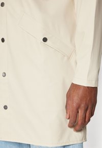 Lys beige vanntett jakke med trykknapplukking, sidelommer og en jevn tekstur. Ermekantene er tilpasset uten synlig utstyr.