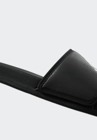 Black slip-on sandal with a textured leather strap, soft fabric lining, and a flat sole. Features a wide, padded upper for comfort.