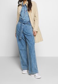 Combinaison en jean à jambes larges, taille ceinturée et col, portée sous un trench-coat beige. Des chaussures de sport blanches complètent la tenue.