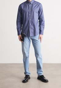 Camicia navy con bottoni e colletto, in tessuto leggero, abbinata a jeans azzurri chiari e scarpe nere stringate. Tiene un portafoglio nero.