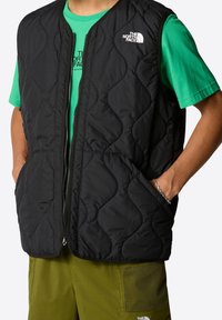 Black quilted vest with a zipper front, featuring two side pockets and The North Face logo. Worn over a green T-shirt and olive shorts.