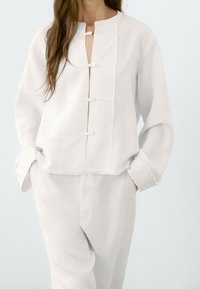 White linen blouse with a round collar, front button closure, and textured panel detailing. Loose fit with long sleeves.