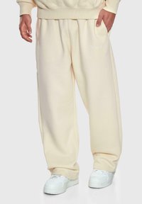 Mann trägt cremefarbene, weit geschnittene Sweatpants und weiße Sneakers, Hand in der Tasche, sichtbar von der Taille bis zu den Füßen.