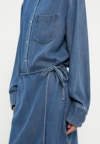 Φόρεμα πουκάμισο από τζιν με μακριά μανίκια, μια τσέπη μπροστά και ζώνη στη μέση. Το ύφασμα είναι μεσαίου μπλε χρώματος με υφή και κλείνει με κουμπιά.