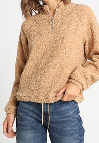 Pull polaire beige avec col à fermeture éclair partielle. Matière texturée et douce. Comprend des poignets élastiques et un ourlet avec cordon de serrage. Porté avec un jean bleu.