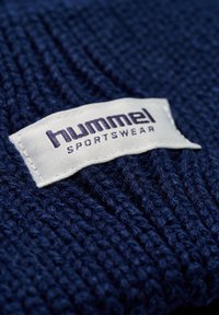 Marinblått stickat tyg med en vit etikett med texten "hummel" och "SPORTSWEAR" i mörkblått, broderad längs kanten.