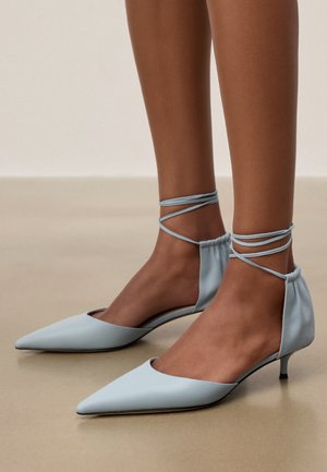 Kurze braune Beine mit hellblauen spitz zulaufenden Kitten-Heels mit Knöchelriemen, die auf einem beigen Boden stehen.