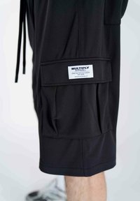 Pantaloni cargo neri realizzati in tessuto mesh, dotati di una grande tasca laterale con etichetta. Il design include cordini e una vestibilità rilassata.