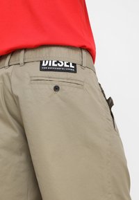 Shorts color khaki con una texture liscia, dotati di una tasca posteriore con bottone e un'etichetta nera con la scritta "DIESEL PER UNA VITA DI SUCCESSO."