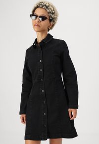 Crna traper haljina s preklopom na dugmad, ovratnikom, dugim rukavima i strukiranim strukom s lepršavom suknjom. Glatka tekstura i minimalistički dizajn.