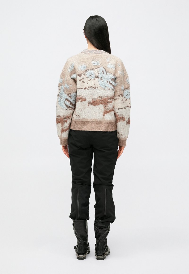 Maglione beige con motivi astratti blu e marroni, polsini e vita a coste, abbinato a pantaloni neri e stivali neri al ginocchio.