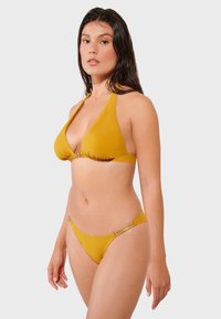 Žuti bikini set koji sadrži trokutasti gornji dio s dekorativnim metalnim detaljem i donji dio s niskim strukom s perlicama. Glatka tekstura.