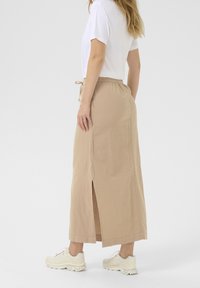 Jupe maxi beige avec fente latérale, en tissu léger, dotée d'une taille élastique avec cordon de serrage, associée à un t-shirt blanc et des baskets.