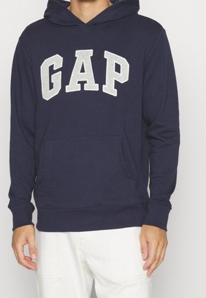 Persona che indossa un hoodie blu navy con le lettere grigie "GAP" sul davanti e pantaloni bianchi, in piedi su uno sfondo semplice.