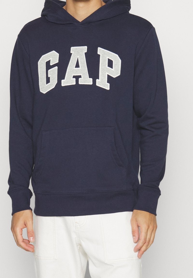 Person som bär en marinblå huvtröja med grå "GAP"-bokstäver på framsidan och vita byxor, stående mot en neutral bakgrund.