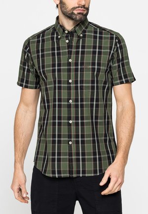 Uomo che indossa una camicia a maniche corte a quadri verde e nero con colletto e tasca frontale, abbinata a pantaloni neri.