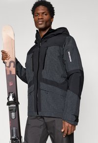 Bărbat îmbrăcat cu o geacă de schi cu carouri negre și gri, ținând o schi vertical cu legături negre pe un fundal simplu.