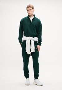 Felpa verde con zip e dettagli bianchi, abbinata a pantaloni jogger verdi. Maglione bianco annodato in vita, indossato con sneakers bianche.