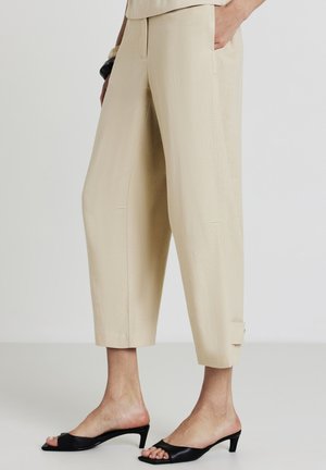 Pantalons beiges raccourcis en matériau doux, avec poches latérales et coupe ajustée. Associés à des mules à talons ouverts noires.